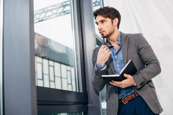 Pensativo hombre de negocios de pie junto a la ventana y la celebración de cuaderno en la oficina - foto de stock