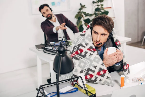 Sick call center operator with blanket on shoulders holding cup and sitting at desk in office — Stock Photo