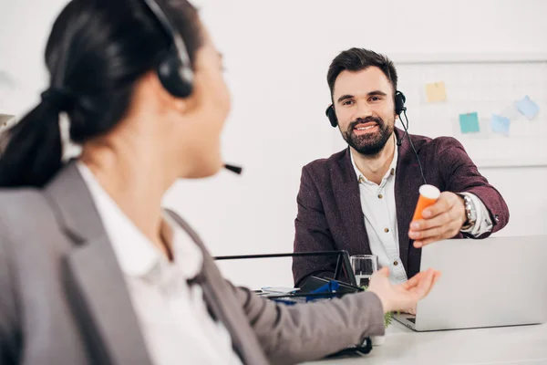 Handsome call center operator smiling and giving bottle with medicine to coworker in office — Stock Photo