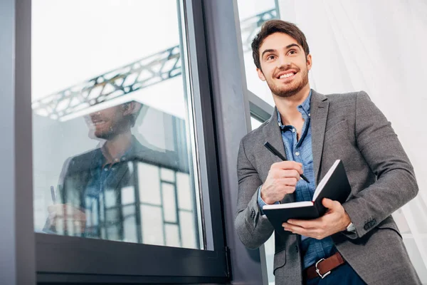 Uomo d'affari sorridente in piedi vicino alla finestra e tenendo aperto il taccuino in ufficio — Stock Photo