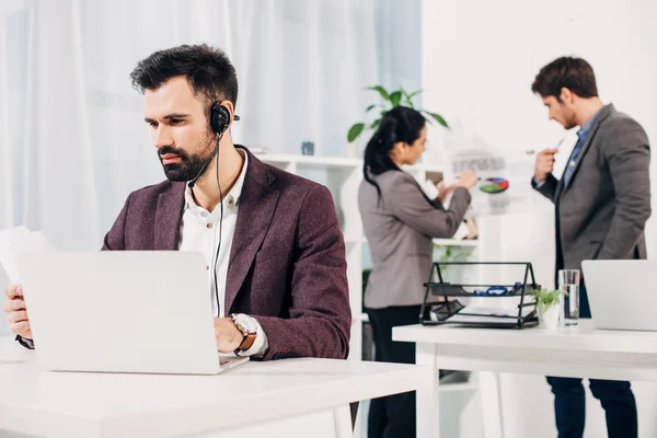 Operador de call center trabalhando no laptop com colegas de trabalho em segundo plano no escritório — Fotografia de Stock