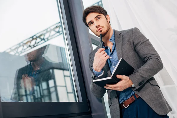 Empresário pensativo em pé à janela com caderno aberto e caneta no escritório — Fotografia de Stock