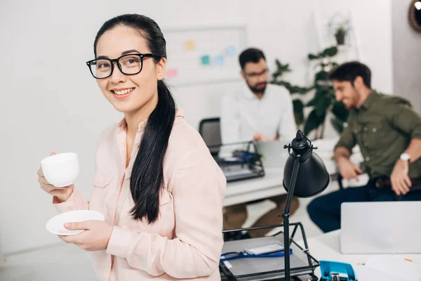Atractivo gerente de oficina sosteniendo taza de café y mirando a la cámara con compañeros de trabajo en el fondo - foto de stock