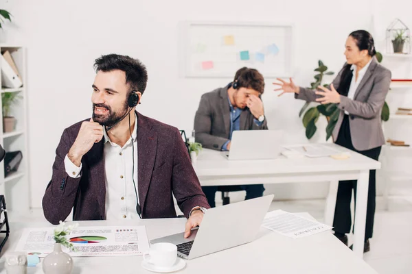 Smiling call center operator sitting at desk with brawling colleagues on background — Stock Photo