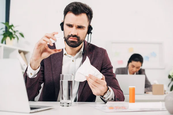 Sick call center operator sitting at desk and holding pill bottle and napkin in office — Stock Photo