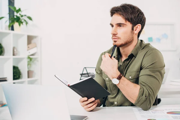 Pensive office manager sitting at desk and holding notebook and pen in office — Stock Photo