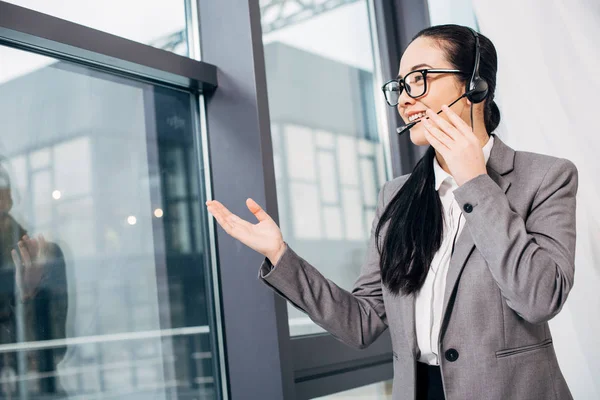 Pretty call center operator touching headset and speaking in microphone by window in office — Stock Photo