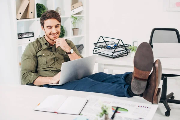 Cheerful businessman sitting on chair with legs on table and looking at laptop in office — Stock Photo