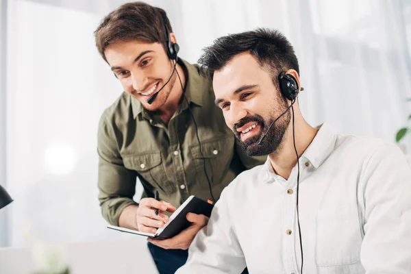 Close up de gerente masculino segurando notebook e caneta perto de colega de trabalho em fones de ouvido no escritório moderno — Fotografia de Stock