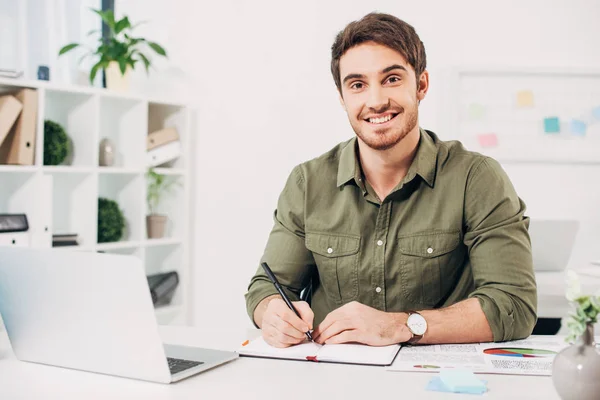Jovem empresário sorrindo e segurando caneta no escritório moderno — Fotografia de Stock