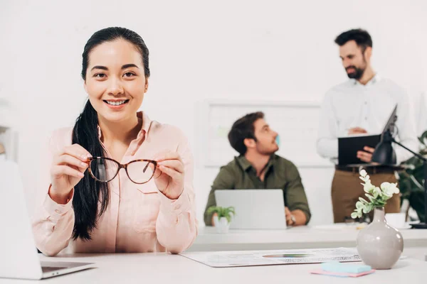 Jovem gerente feminina segurando óculos e sorrindo no escritório — Fotografia de Stock