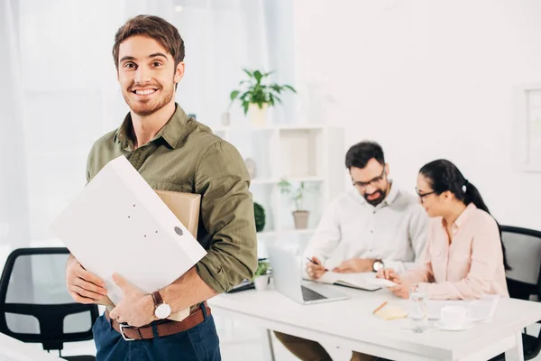 Junger männlicher Büroleiter steht und hält Ordner im Büro mit Mitarbeitern im Hintergrund — Stockfoto