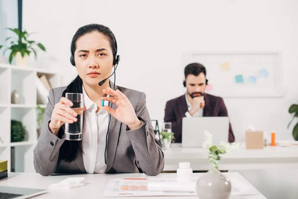 Selective focus of sick female manager holding pill and glass of water in office — Stock Photo
