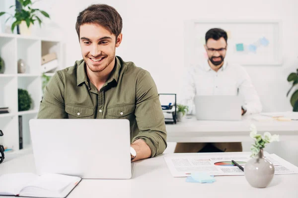 El foco selectivo del manager alegre usando el portátil con el compañero de trabajo en el fondo - foto de stock