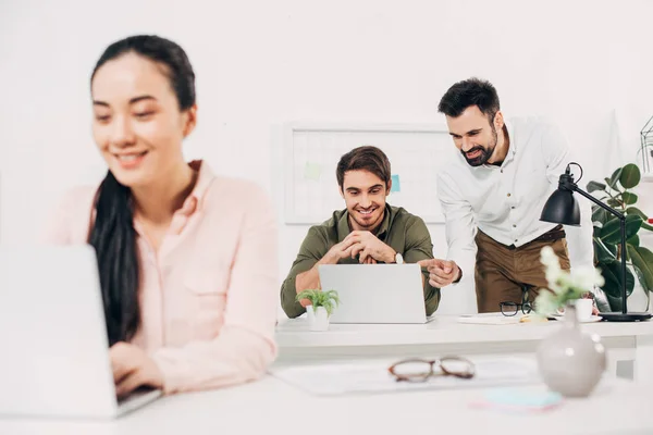 Enfoque selectivo de compañeros de trabajo masculinos mirando a la computadora portátil y sonriendo en la oficina - foto de stock