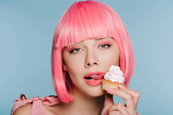 Chica de moda en rosa peluca lamiendo crema de mantequilla de cupcake aislado en azul - foto de stock
