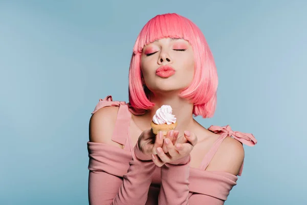 Elegante chica en rosa peluca celebración dulce cupcake y soplado beso aislado en azul - foto de stock