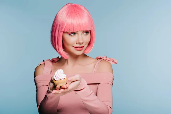 Encantadora chica de peluca rosa sosteniendo sabroso cupcake con crema de mantequilla aislado en azul - foto de stock