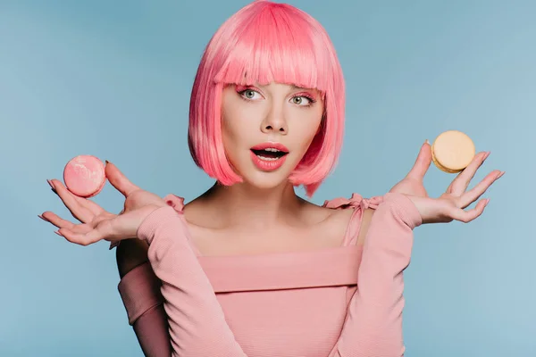 Atractiva chica sorprendida en peluca rosa posando con macarrones aislados en azul - foto de stock