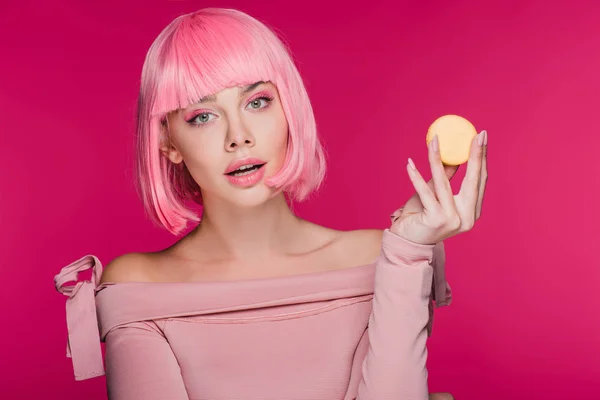 Atractiva chica de moda en peluca rosa posando con macaron amarillo aislado en rosa - foto de stock