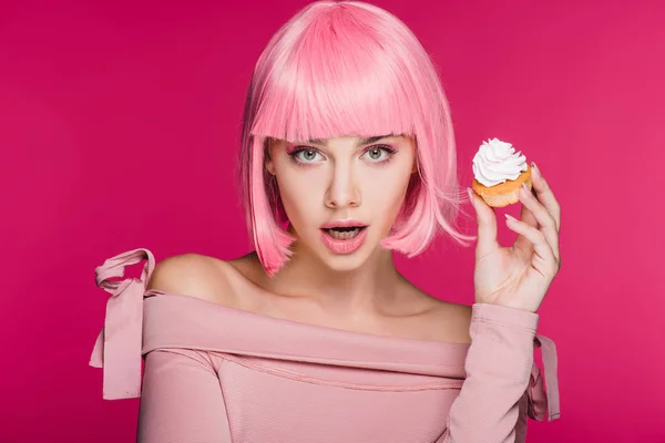 Shocked girl in pink wig holding sweet cupcake isolated on pink — Stock Photo