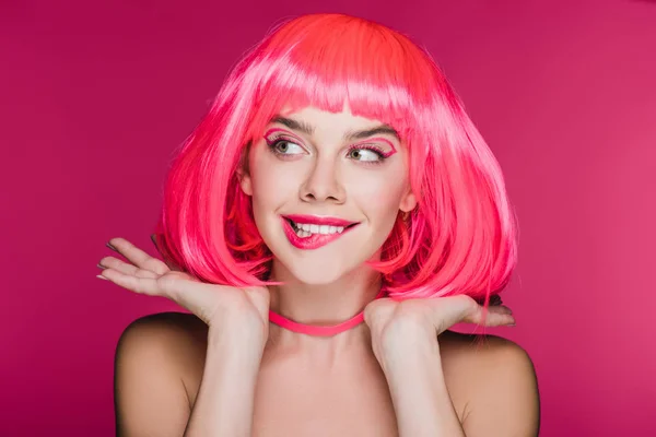 Hermosa chica mordiendo el labio y posando en peluca de neón rosa, aislado en rosa - foto de stock