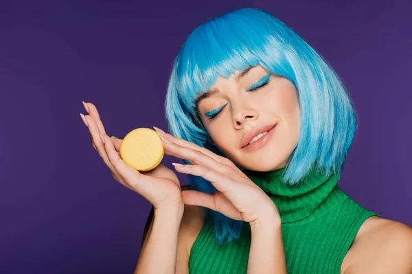 Мрійлива дівчина в синій перуці позує з макароном ізольована на фіолетовому — стокове фото
