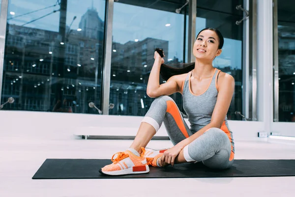 Attractive asian sportswoman sitting on fitness mat and holding hair at modern gym — Stock Photo