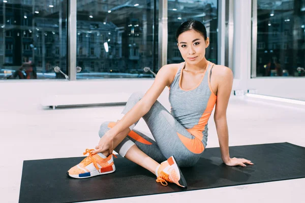Smiling asian girl sitting on fitness mat and looking at camera in modern sports center — Stock Photo