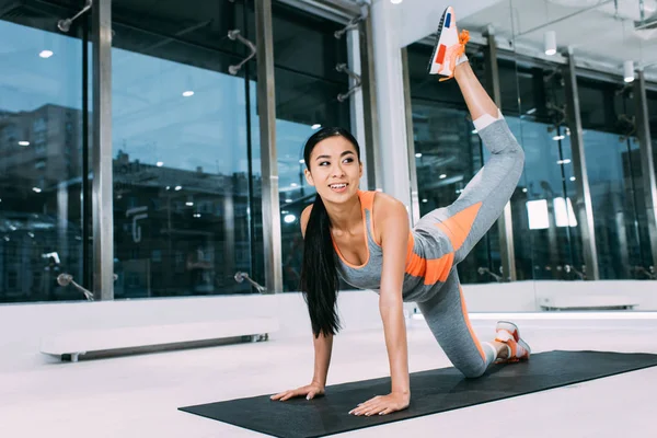 Attractive asian girl smiling and doing aerobic exercise on fitness mat at modern gym — Stock Photo