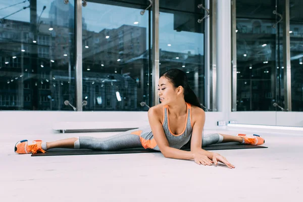 Sonriente asiático chica haciendo split en fitness mat en gimnasio - foto de stock