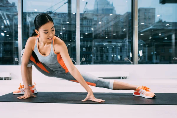 Flexible asiático chica sonriendo y estiramiento pierna en fitness mat en gimnasio - foto de stock