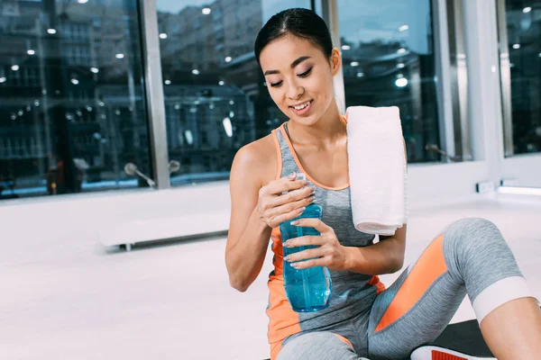 Lächelndes asiatisches Mädchen mit Handtuch auf der Schulter sitzt auf der Fitnessmatte und hält eine Sportflasche mit Wasser in einem modernen Fitnessstudio — Stockfoto