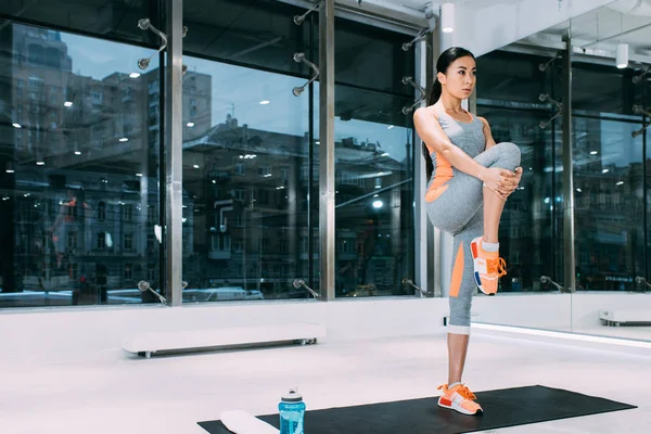 Стройная азиатская девушка, стоящая на коврике для фитнеса и растягивающая ноги в современном спортзале — стоковое фото