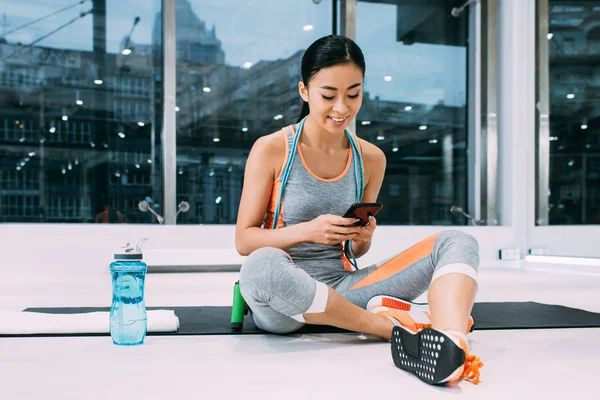 Attractive asian girl sitting on fitness mat and using smartphone in sports center — Stock Photo