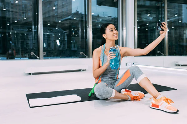 Sorridente ragazza asiatica seduta sul tappeto fitness, con in mano bottiglia sportiva con acqua e scattare selfie in palestra — Foto stock