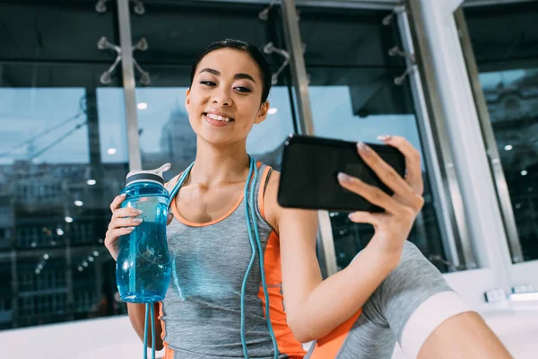 Close up vista de sorrir asiático menina segurando esportes garrafa com água e tomando selfie no ginásio — Fotografia de Stock