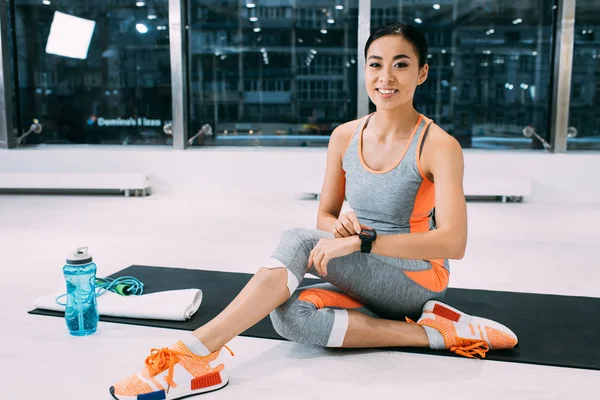 Sonriente asiático sportswoman sentado en fitness mat y tocando fitness tracker en gimnasio - foto de stock