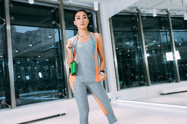Attrayant asiatique fille avec saut de corde sur les épaules regardant caméra à la salle de gym — Photo de stock