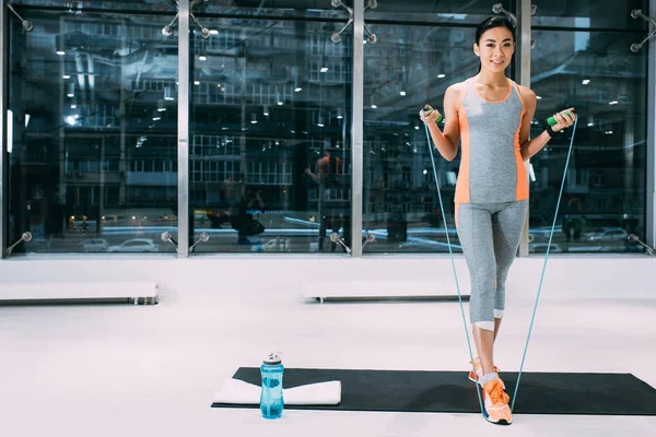 Усміхнена азіатська спортсменка з пропусканням мотузки, що стоїть на фітнес-матусі і дивиться на камеру в спортзалі — стокове фото