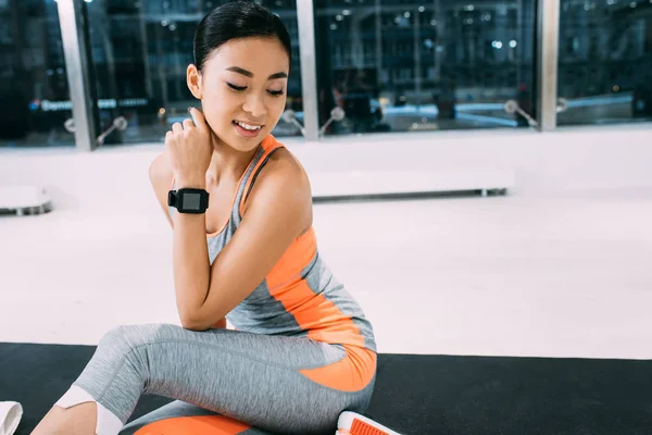 Улыбающаяся азиатская спортсменка с фитнес-трекером, сидящая на коврике в спортзале — стоковое фото