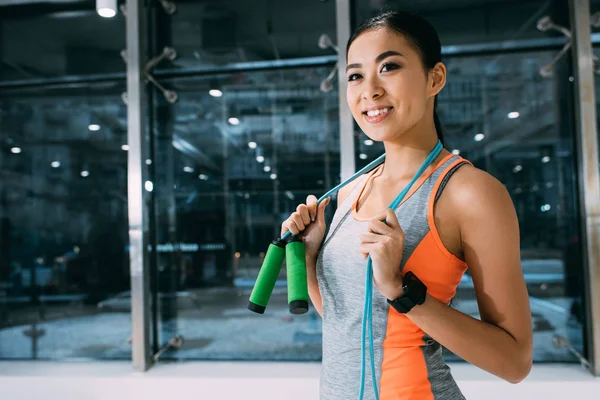 Sonriente asiático sportswoman holding saltar cuerda en hombros en gimnasio - foto de stock