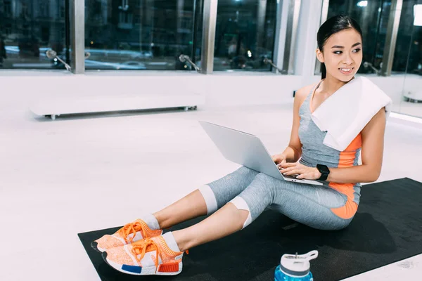 Jovem asiático esportista com toalha no ombros sentado no tapete de fitness, digitando no teclado do laptop e sorrindo no ginásio — Fotografia de Stock