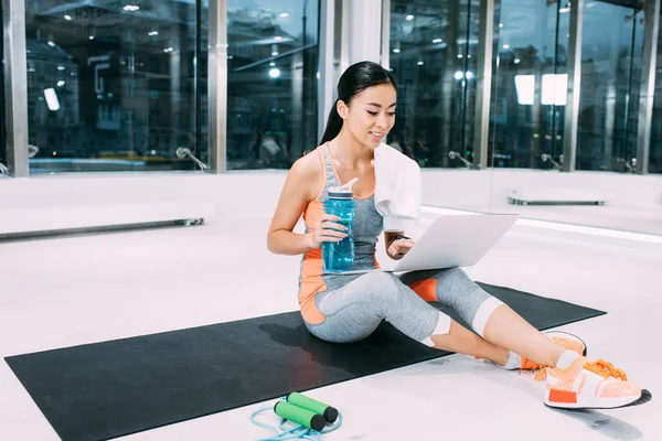 Sorridente sportiva asiatica seduta sul tappeto fitness, digitando sulla tastiera del computer portatile e tenendo bottiglia sportiva in palestra — Foto stock