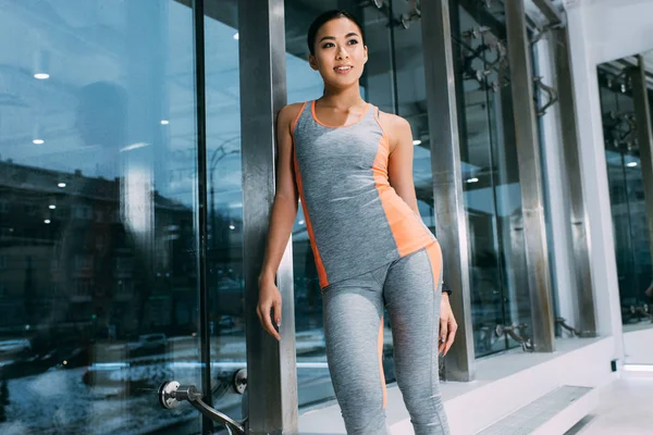 Привлекательная азиатка в спортивной одежде, стоящая у окна и улыбающаяся в современном спортзале — стоковое фото