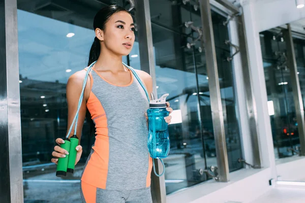 Beautiful asian sportswoman holding sports bottle with water and skipping rope at gym — Stock Photo