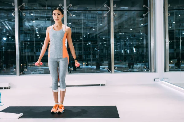 Attrayant asiatique sportive debout sur tapis de fitness et faire de l'exercice avec haltères à la salle de gym — Photo de stock