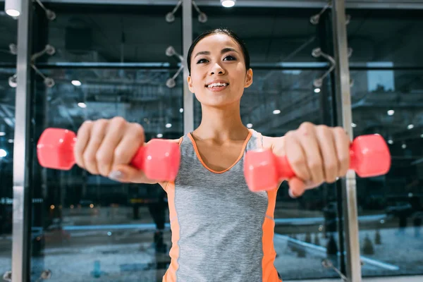 Close up view of attractive asian sportswoman smiling and doing exercise with dumbbells at gym — Stock Photo
