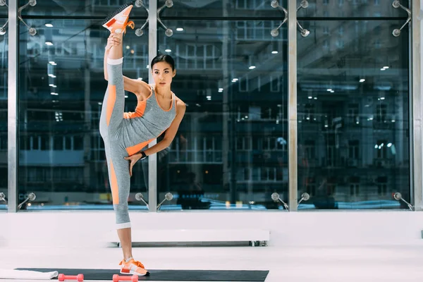 Flexible asiático sportswoman con pierna arriba de pie en fitness mat en gimnasio - foto de stock