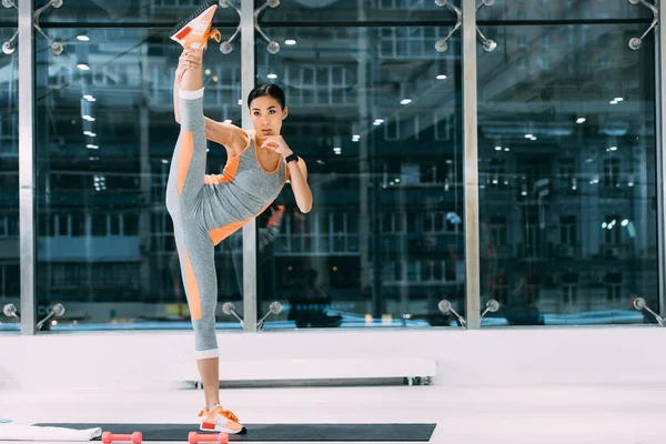 Atlético asiático chica en sportswear con pierna arriba de pie en fitness mat en gimnasio - foto de stock